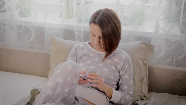 A young girl with dark hair sitting on a bed in her pajamas with stars in your day off and overwritten with someone on the phone. Woman happy and responds to messages with a smile