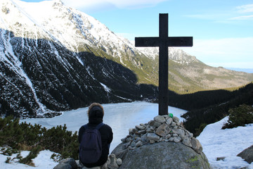 A young man sits near a large cross on a background of mountains.