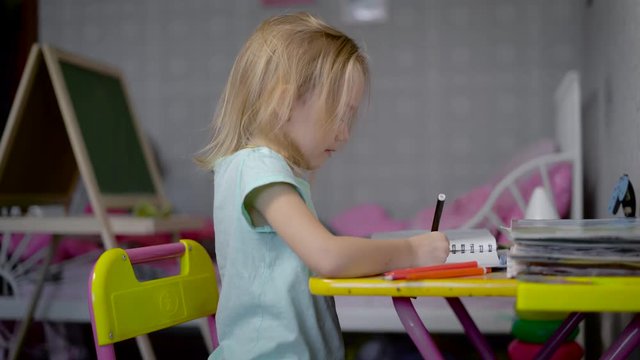 Little girl is drawing the picture with pencils in studio sitting at the table. Small child is learning to paint with soft-tip pens of different colours. Cute kid crayoning the pattern with pleasure.