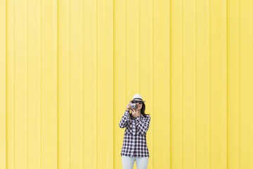 Happy beautiful woman talking a selfie with a vintage camera over yellow background. Wearing hat and sunglasses.