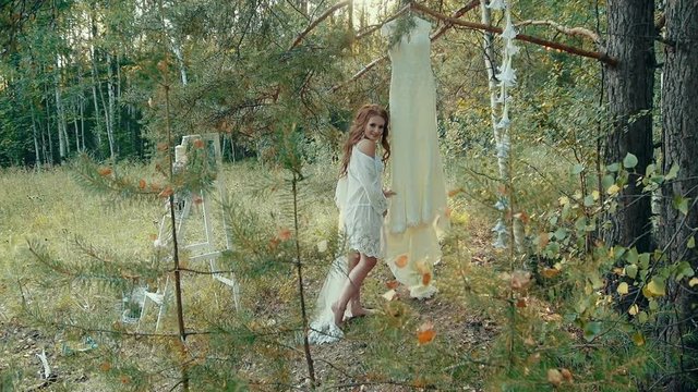 Young caucasian woman in white sexy lingerie with lace long veil on her head touches her wedding dress hanging on the branch of a pine in the autumn forest in the afternoon.