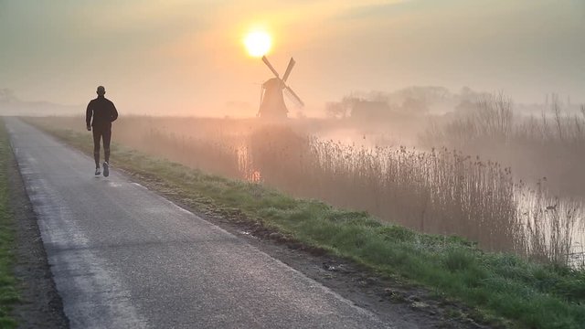 Man running near a windmill in the Dutch countryside during a foggy, spring sunrise.