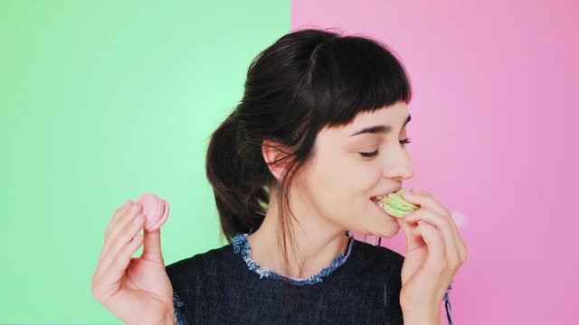 Beauty smiling young brunette isolated on splitted pistachio and pale rose red background holds two tasty cookies in her hands and makes difficult choice, then bite right macaron in pastel green color