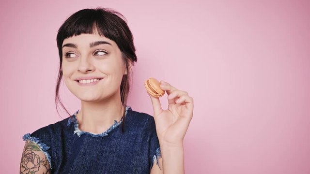 Young fresh and beautiful brunette girl isolated on side on pale pink background smiles, thinks about something, looks on sides and holds small macaron cookie in her hand, wearing denim dress