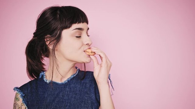 Young fresh and beautiful brunette girl isolated on side on pale pink background smiles, thinks about something, looks on small macaron cookie in her hand and bites it, wearing denim dress
