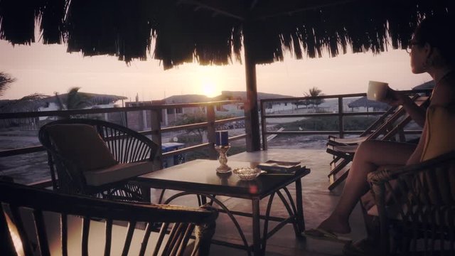 Hispanic woman drinking coffee under a hut near the beach during a marvelous sunset. 4k