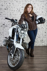 Attractive woman in vintage outfit holds white helmet, standing near motorcycle, brick wall background