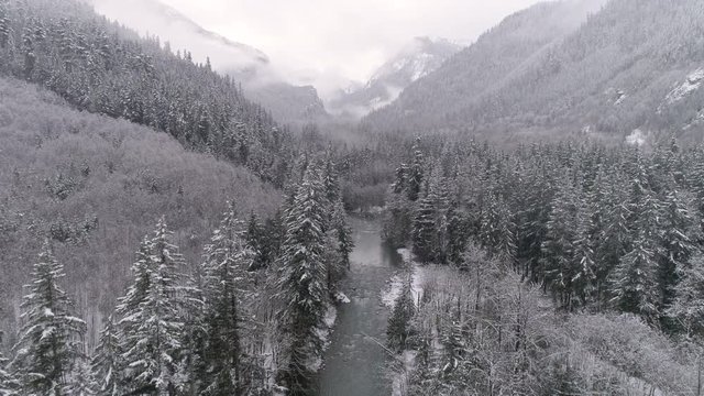 Aerial of Frozen Winter Landscape in Mountain Forest Valley with Snow Flakes Falling on Ice Cold River