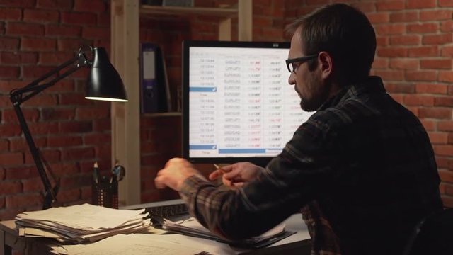 back view of Caucasian male in glasses sitting in front of monitr and foraging among papersin slowmotion