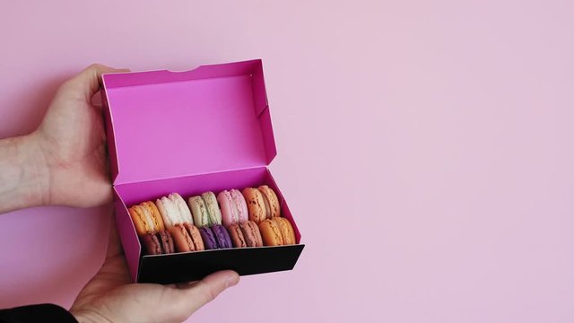 Hands moves in small gift box wtih multicolored macarons and open it, isolated on pale rose background