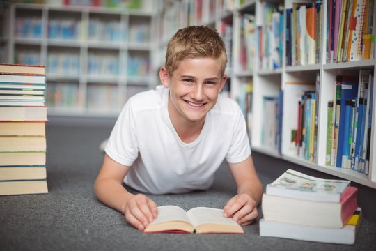 Portrait of happy schoolboy lying on floor with books in library