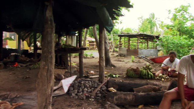 Samoan man scraping coconut meat and woman peeling plantains near cooking fire