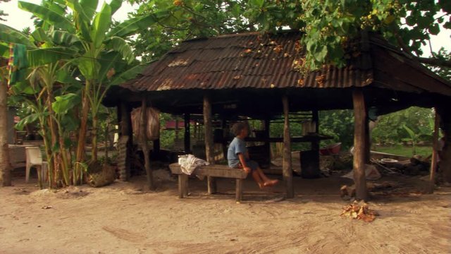 Samoan child seated under metal roof of open-sided hut while family works nearby