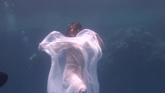 Underwater model young girl free diver in white transparent veil in Red Sea. Filming a movie at camera. Extreme sport in marine landscape, coral reefs, ocean inhabitants.