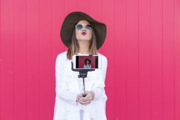 Happy beautiful woman taking a selfie with smartphone over red background. Wearing hat and sunglasses and using selfie stick