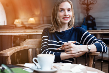 A smile young woman dreams seat in a cafe with cup of coffee with wooden pencil in hand. Concept of planning personal traning schedule. Looking at camera.