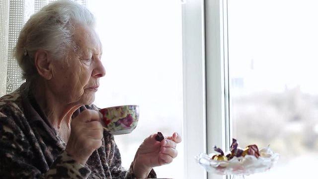 Sad old woman looking out the window and drinking tea. Close up