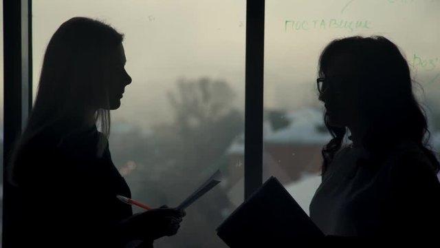 By the window there are two silhouettes of women with papers in hand. They lead talking. Staff in the office talking and holding in their hands paper and pens. Behind the glass the evening winter