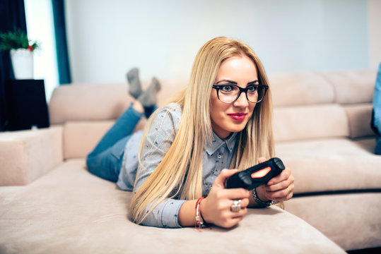 Blonde girlfriend enjoying video games on console while laying on living room sofa