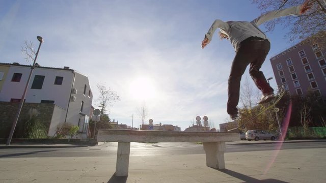 SLOW MOTION CLOSE UP, LOW ANGLE: Pro skateboarder jumping, sliding on concrete bench on the street against the sun. Skater skateboarding & doing grinding trick with skateboard in city residential area