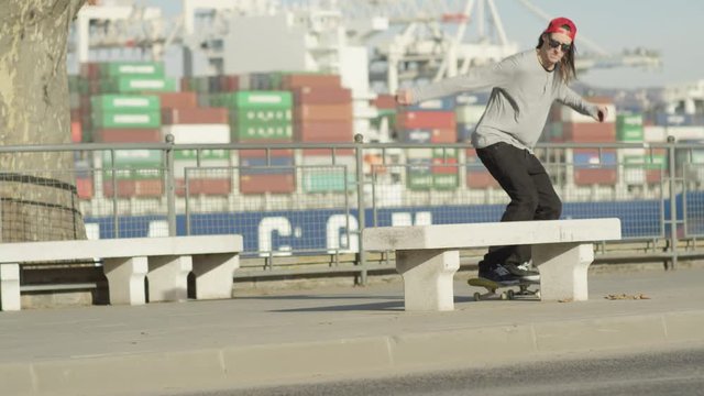 SLOW MOTION CLOSE UP: Extreme pro young skateboarder riding skateboard and jumping in front of seaport and loaded container ship. Skater doing grind trick with skateboard sliding on concrete bench