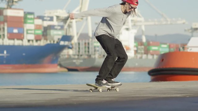 SLOW MOTION CLOSE UP: Pro skateboarder skateboarding and jumping heelflip trick riding along the concrete ocean coast on sunny day with freighter ships anchored in the seaport harbor in the background