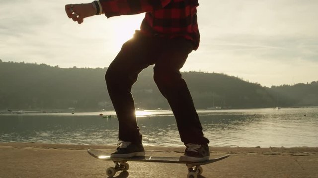 SLOW MOTION, CLOSE UP: Unrecognizable skateboarder skateboarding and jumping 360 flip trick on promenade along the ocean at golden light sunset. Skateboarder riding skateboard at sunrise at seaside