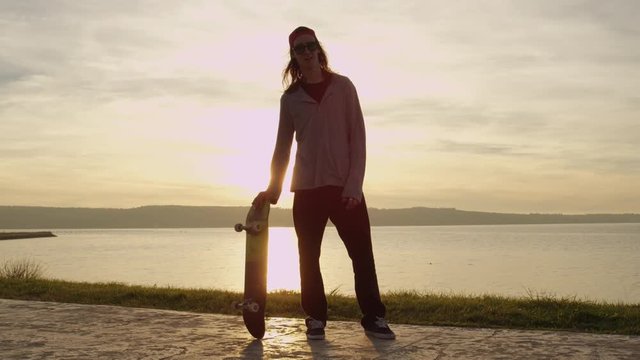 SLOW MOTION, CLOSE UP: Young skateboarder having fun playing with skateboard spinning and revolving it at golden light sunset. Skater standing on the concrete shoreline at the end of skateboarding day