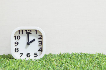Closeup white clock for decorate in 2 o'clock on green artificial grass floor and cream wallpaper textured background with copy space