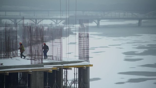 workers walking on the construction site and check. Man wearing in orange coveralls and hard hat. Urban view on the city with snow ice on river and bridge.
