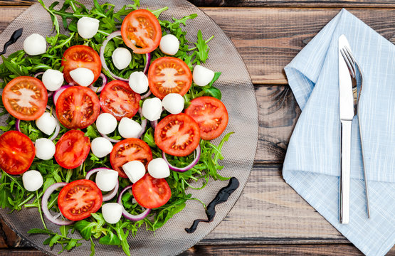 Organic vegetables salad: cherry tomatoes, mozzarella cheese, rocket, red onion on clear plate over dark rustic wooden table. Traditional italian salad ingredients. Mediterranean food. Top view. 