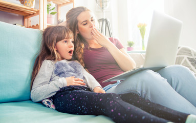 Mother yawning while excited daughter watching movie on laptop