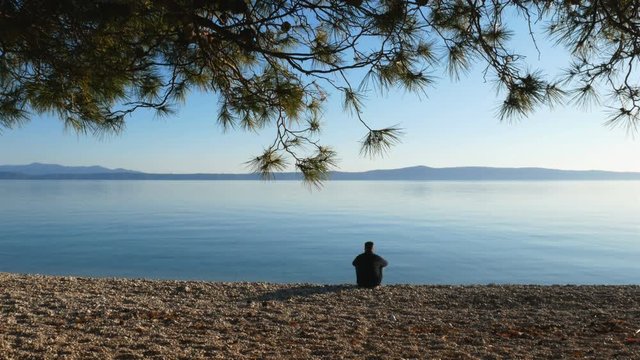 Young man sitting alone on the beach and looking at calm blue sea on sunny day