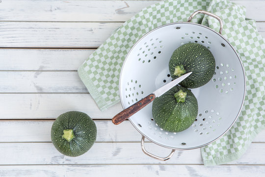 Runde Zucchini in einem Lochsieb und einem Messer auf einem alten weissen Holzuntergrund