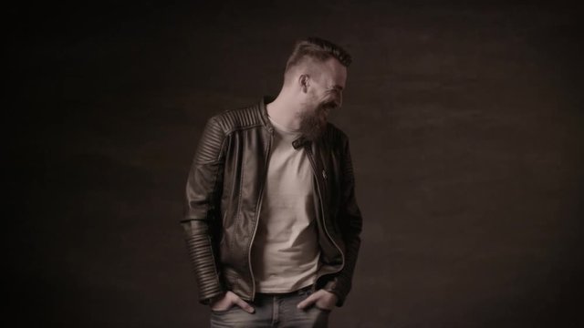 Handsome man with a beard in a black leather jacket posing in the studio on a black background.A man with a beard laughs