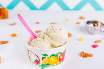 Close up fruit Ice-cream in a paper cup on the white background with colorful candies, pieces of waffles, ice cream spoon and empty ice cream cone. Selective focus.