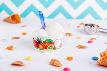Close up Vanilla Ice-cream in a paper cup on the white background with colorful candies, pieces of waffles, ice cream spoon and empty ice cream cone. Selective focus.