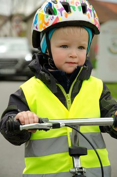 Little child learns to ride a bike on the road. There is a car in the background. Boy is marked by yellow reflective vest and helmet because of visibility and safety.