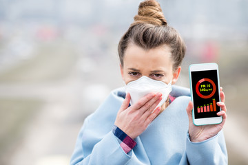 Woman in protective mask holding smart phone with air polution measurement of PM10 outdoors - obrazy, fototapety, plakaty