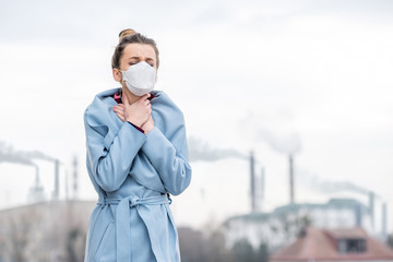 Young woman in protective mask suffering from air pollution from big manufacturing in the city