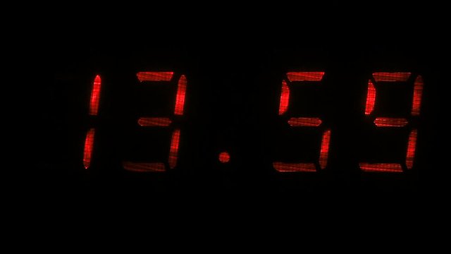 Digital clock with fluorescent display shows the time of 13 hours 59 minutes to 14 hours 00 minutes in the red on a black background