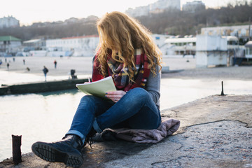 Young woman with red hair curls sitting and drawing with pen at the seacoast