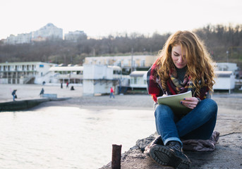 Young woman with red hair curls sitting and drawing with pen at the seacoast