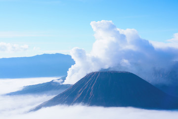 White smoke coming out of volcanoes surrounded by white clouds of mist and a clear blue sky seen at a distance in the afternoon at National Park in East Java, Indonesia.