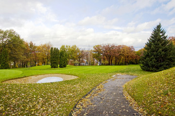 Colors of the fall at Midwest USA. Fallen leaves on a green lawn. Autumn landscape with path and puddle. Cloudy sky over golf course and colorful trees around. Horizontal shot.