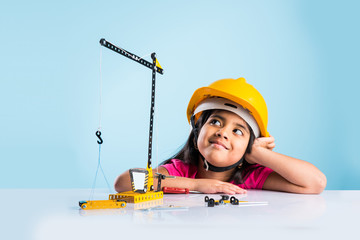 cute indian baby girl playing with toy crane wearing yellow construction hat or hard hat, childhood...