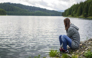 Fototapeta na wymiar Beautiful young woman relaxing by the mountain lake sitting on the edge of lakeshore looking aside. Enjoing nature