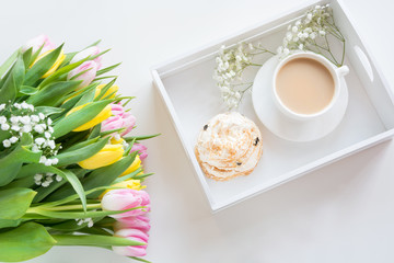 Morning breakfast in spring with a cup of black coffee with milk and pastries in the pastel colors, a bouquet of fresh yellow and pink tulips on a white background. Top view.