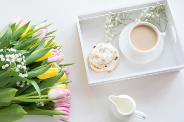 Morning breakfast in spring with a cup of black coffee with milk and pastries in the pastel colors, a bouquet of fresh yellow and pink tulips on a white background. Top view.