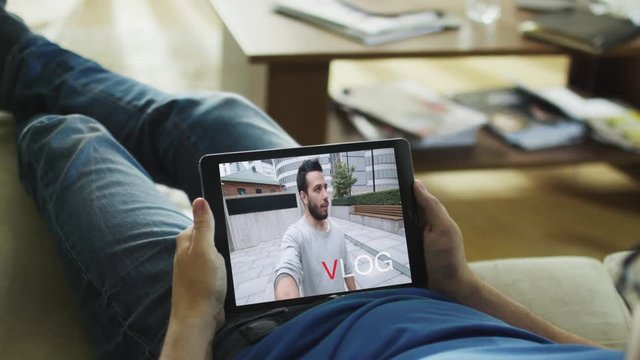 Casual Man Lying on His Couch Watching Fashionable Video Blogg on His Tablet Computer. Inscription "Vlog" appears on the Screen. Shot on RED Epic Cinema Camera in 4K (UHD).